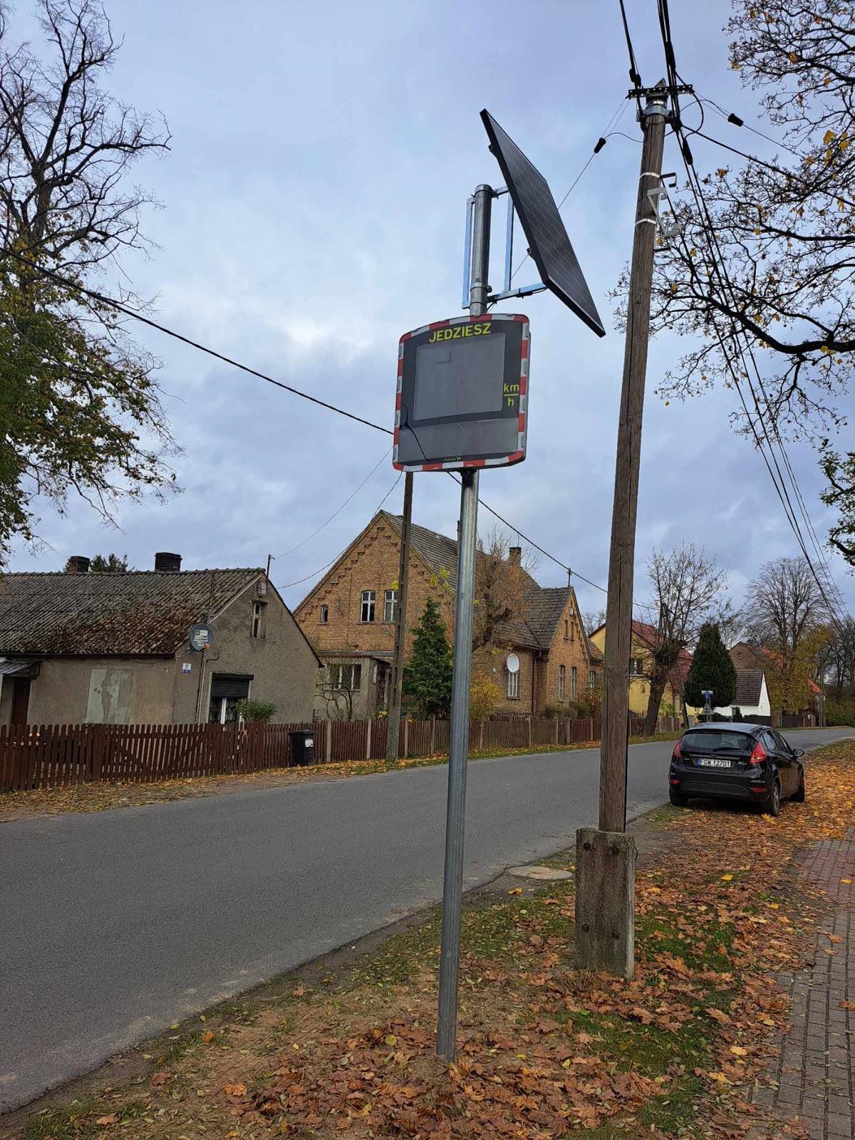 Solarny radarowy wyświetlacz prędkości z napisem „JEDZIESZ” na słupie przy wiejskiej drodze, jesienią