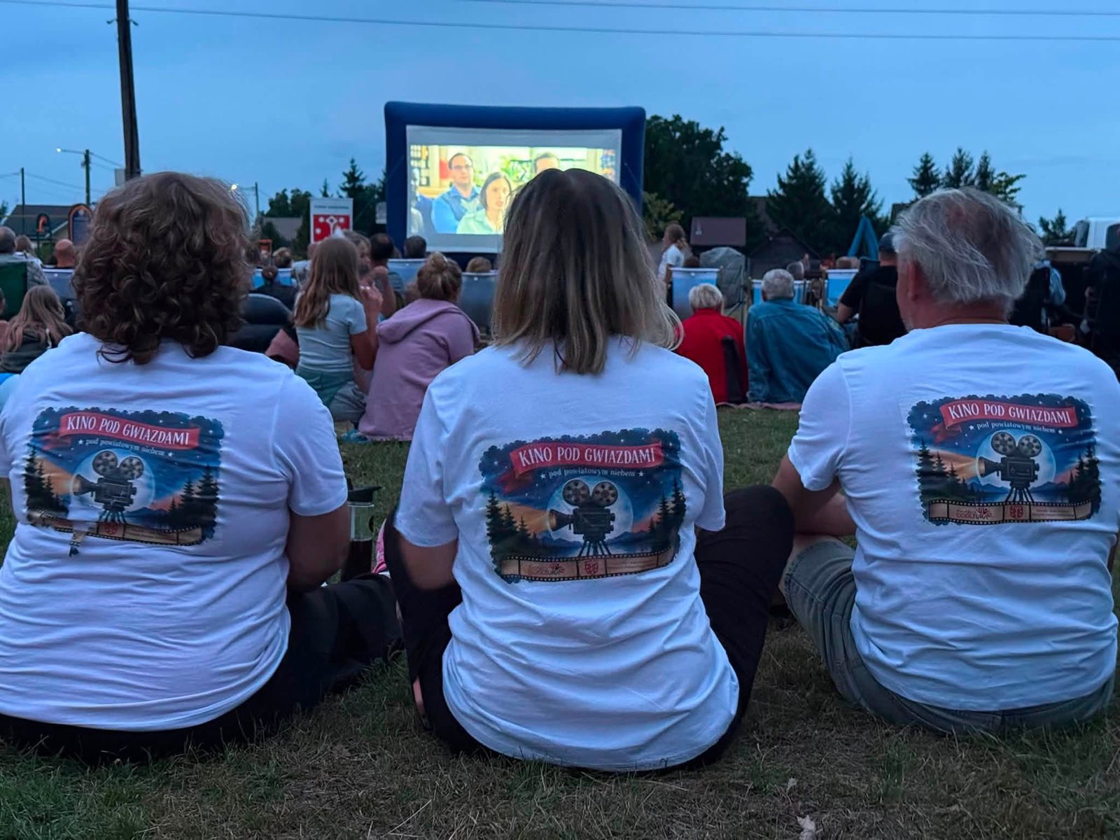 Trzy osoby w koszulkach „Kino pod Gwiazdami” siedzą na trawie przed dmuchanym ekranem, w tle publiczność o zmierzchu